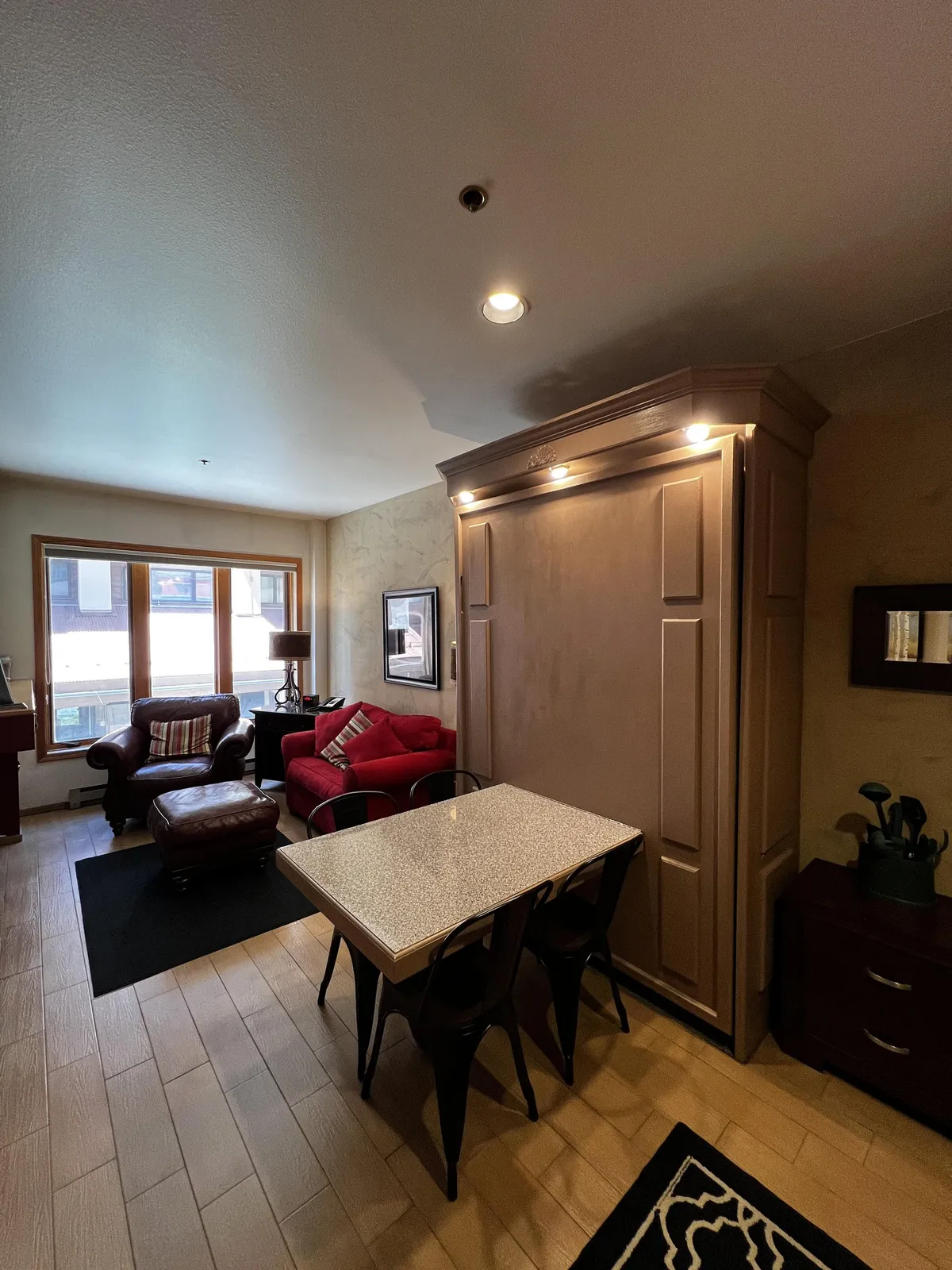 This wide-angle shot displays the main studio space with a Murphy bed, dining table, and large windows that likely frame mountain views. The red armchair and the overall open layout highlight how the space can shift between sleeping, lounging, and dining, emphasizing the studio's clever design and flexibility.