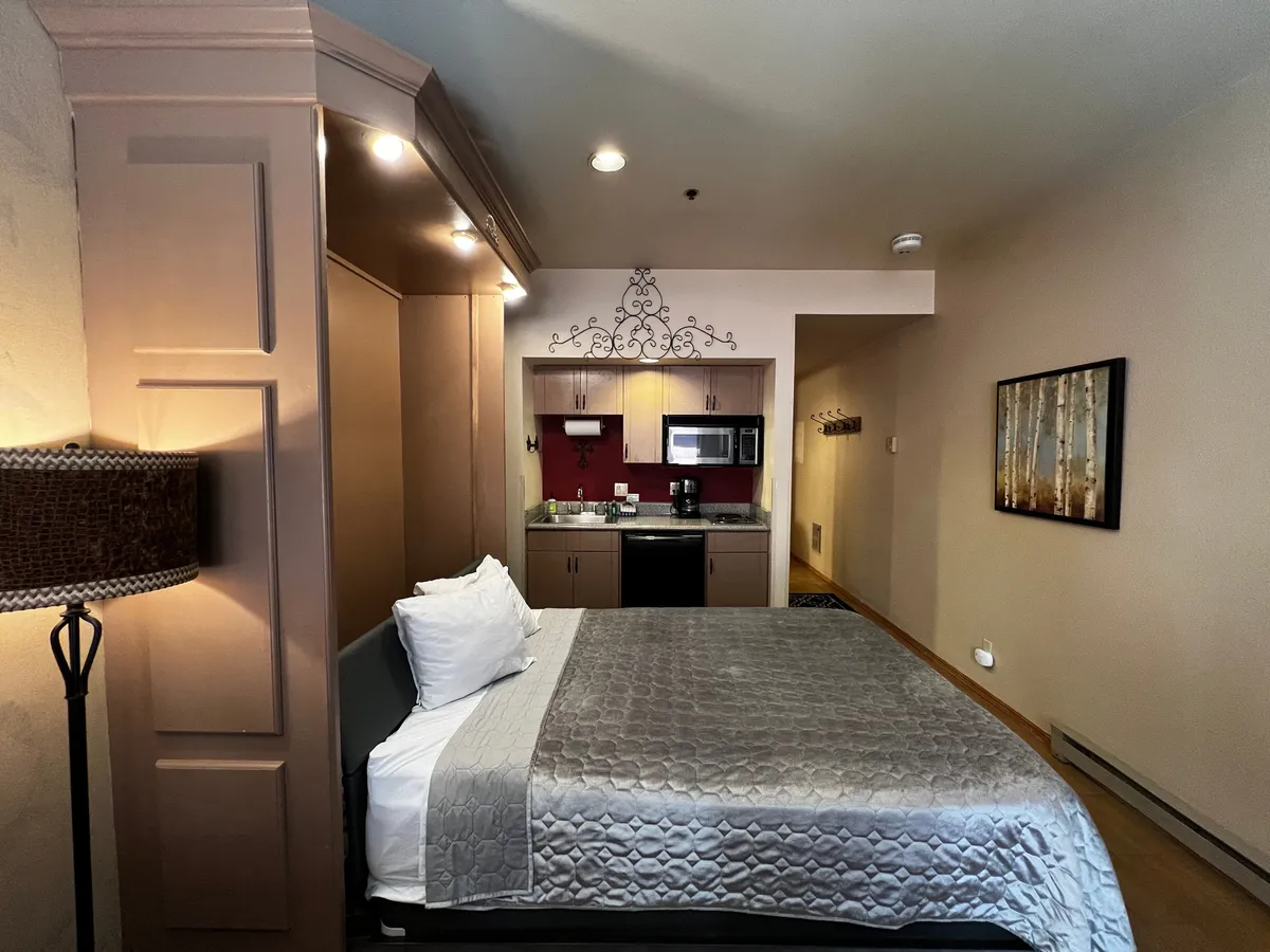 This image focuses on the cozy sleeping area with the queen-sized bed, adjacent kitchenette, warm lighting, and tasteful artwork. It highlights an inviting, homely atmosphere ideal for morning routines or winding down at night. The arrangement demonstrates how the sleeping and living spaces blend seamlessly.