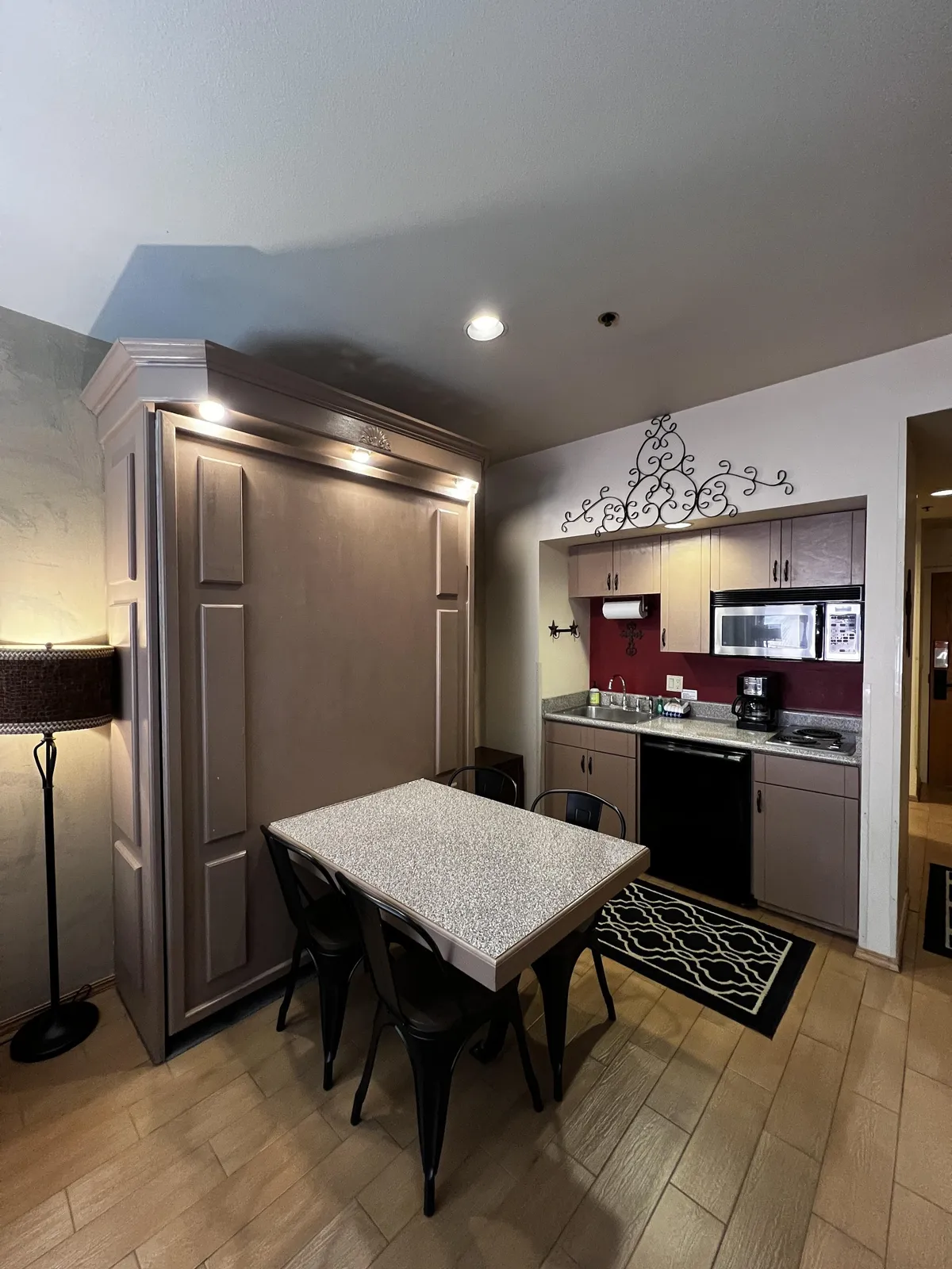 This image features a compact dining table adjacent to kitchen cabinetry, with a coffee maker prominently visible. Decorative wall art adds a touch of warmth, and the space is clearly designed to invite family meals, cooking together, and social gatherings. The angle shows the heart of the kitchenette and dining area working together as an inviting hub.