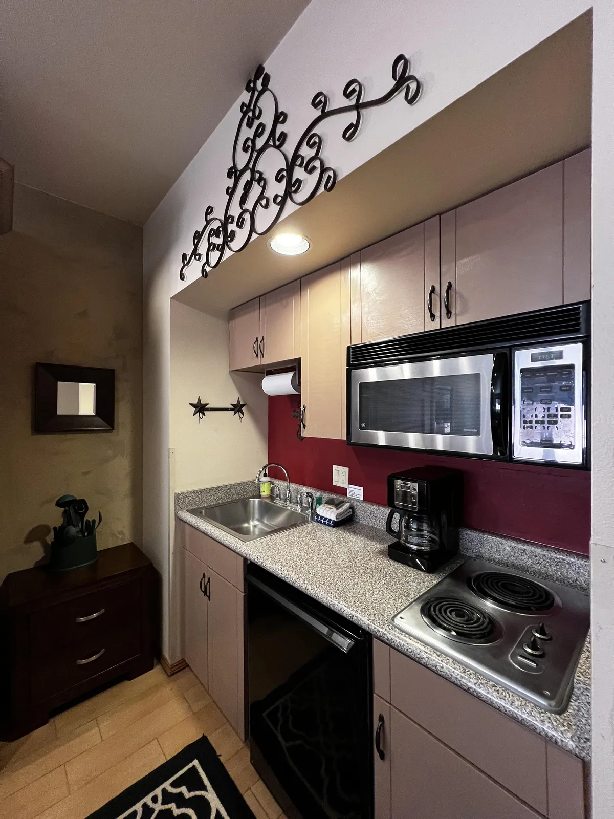 The focus is on a granite countertop, black appliances, decorative wall art, and various kitchen utensils. The intimate setup suggests spaces ideal for casual brunches, baking sessions, and socializing while preparing food.