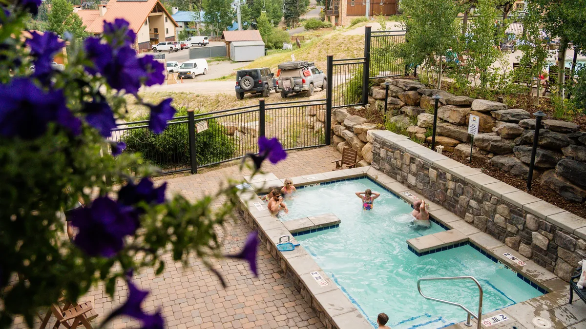 An inviting exterior shot showing a hot tub surrounded by greenery and flowering plants, with people swimming and socializing in the background. The bright and casual mood suggests the lively community atmosphere at the base of the slopes.