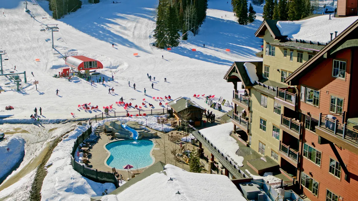 This exterior shot showcases a heated outdoor pool adjacent to the hotel, with skiers and snowboarders nearby and the majestic ski slopes in the background. The scene is vibrant and energetic, framing the pool as both a relaxing and social hub after outdoor adventures.