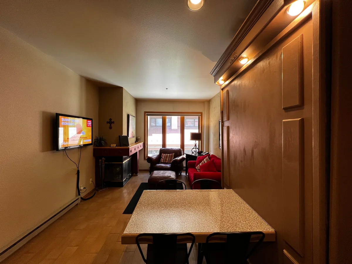 This image captures the main studio living area featuring a cozy red couch, a wood-burning fireplace, a TV, and a dining table. The fireplace is lit, creating a warm, inviting atmosphere perfect for relaxing after a day on the slopes or curling up with a book as snow falls outside. The space appears open and multifunctional, reflecting both comfort and utility.