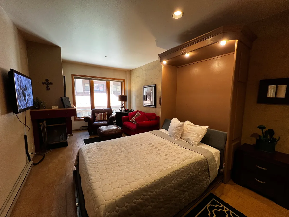 The image shows a cozy condo interior with a neatly made queen-sized Murphy bed, a plush seating area facing a flat-screen television, and large windows letting in natural light and offering glimpses of the surrounding mountain scenery. The ambiance is warm and inviting, perfect for relaxing with a book, enjoying a movie, or sipping coffee while taking in the views.