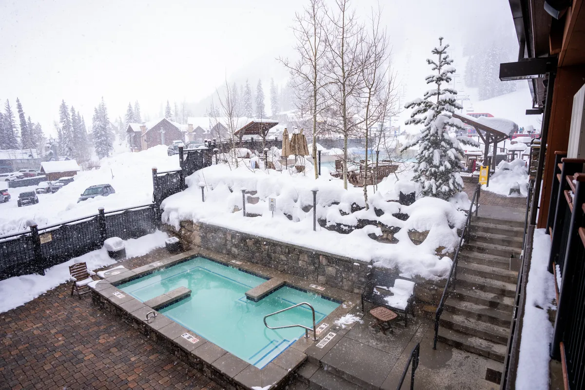 This image shows an inviting outdoor hot tub surrounded by a snow-covered landscape and tall trees, with a staircase leading up to the steaming water. The scene is tranquil, presenting the hot tub as a cozy, luxurious spot for relaxation while enjoying the wintry outdoors. The nighttime atmosphere is perfect for stargazing or unwinding after a day of activity.